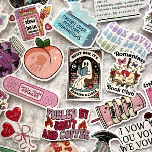 May include: A collection of colorful stickers with various designs and text. Stickers include a peach, a ghost reading a book, a matchbox with "slow burn romance", and a band-aid with "small town romances". Other stickers say "Crying Over Fictional Characters" and "Fueled by Smut and Coffee".
