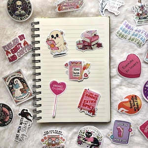 May include: A collection of colorful stickers with book-related themes. Designs include a ghost reading, stacks of books, a matchbook with the text "slow burn romance," and a heart-shaped lollipop that says "Enemies to lovers." Other stickers say "Book Club."