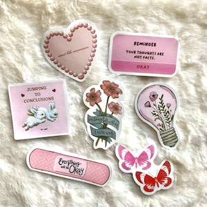 May include: A set of pink and white stickers with various designs, including a heart with the text "sweet like cinnamon", a lightbulb with flowers, a butterfly, a band-aid with the text "Everything will be Okay", a bunny with the text "Jumping to conclusions", and a flower with the text "im dang bollerthan I ever was".