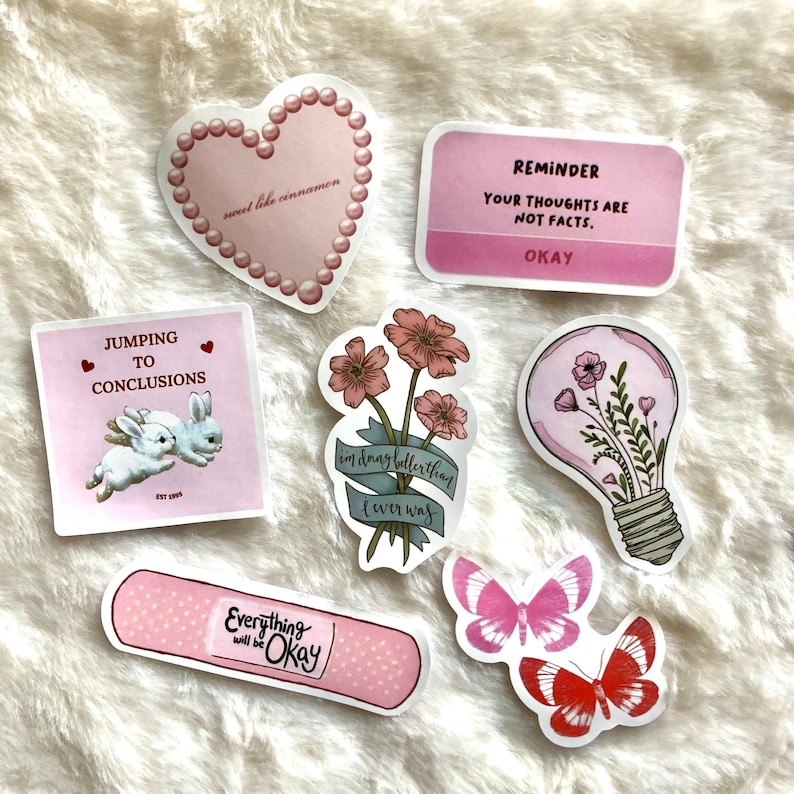 May include: A set of pink and white stickers with various designs, including a heart with the text "sweet like cinnamon", a lightbulb with flowers, a butterfly, a band-aid with the text "Everything will be Okay", a bunny with the text "Jumping to conclusions", and a flower with the text "im dang bollerthan I ever was".