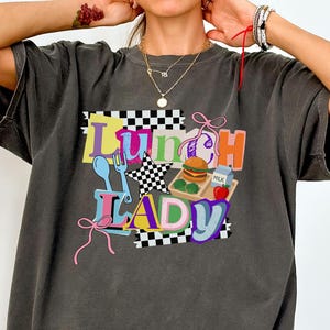 May include: Charcoal grey t-shirt featuring the words "Lunch Lady" in a vibrant, retro font. The design incorporates a checkerboard, a burger, an apple, and a milk carton. The shirt has a loose, comfortable fit.