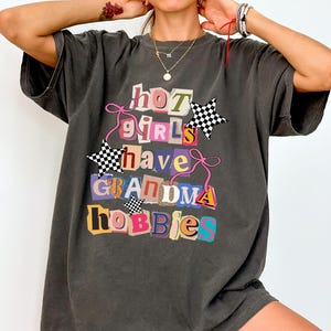 May include: Dark gray oversized t-shirt with the text "hot girls have grandma hobbies" in a colorful, cut-out style. The design includes two checkered star accents and pink string details. The shirt is a relaxed fit.