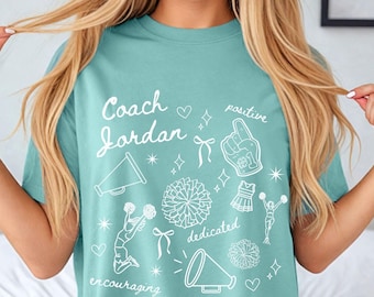 May include: A teal blue t-shirt with a white design that says "Coach Jordan" and includes cheerleading-themed illustrations. The words "positive", "dedicated", and "encouraging" are also included in the design. The shirt is labeled "Chalky Mint".
