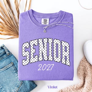 Peut inclure: Un sweat-shirt violet Comfort Colors avec le mot "SENIOR" en lettres blanches &agrave; pois noirs, et l'ann&eacute;e "2027" en dessous. Un collier argent&eacute; avec un pendentif soleil repose sur le v&ecirc;tement. Le mot "Violet" est en bas.