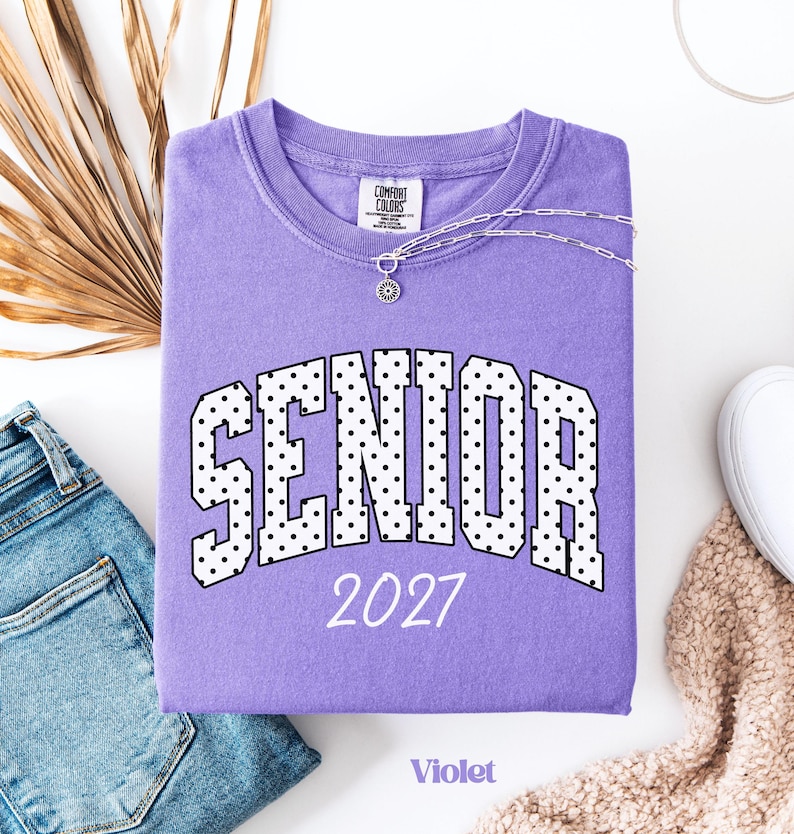 Peut inclure: Un sweat-shirt violet Comfort Colors avec le mot "SENIOR" en lettres blanches &agrave; pois noirs, et l'ann&eacute;e "2027" en dessous. Un collier argent&eacute; avec un pendentif soleil repose sur le v&ecirc;tement. Le mot "Violet" est en bas.