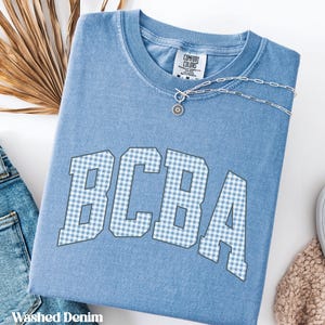 Pode incluir: Uma t-shirt azul de manga curta com acabamento lavado, apresentando o texto BCBA num padrão de xadrez vichy azul claro e branco. A t-shirt tem um decote redondo com uma etiqueta Comfort Colors e uma corrente prateada sobre a gola.