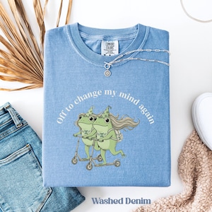 May include: Light blue t-shirt with the text "Off to change my mind again" and a graphic of two green frogs on scooters. The shirt is folded and has a Comfort Colors tag. The words "Washed Denim" are printed at the bottom.