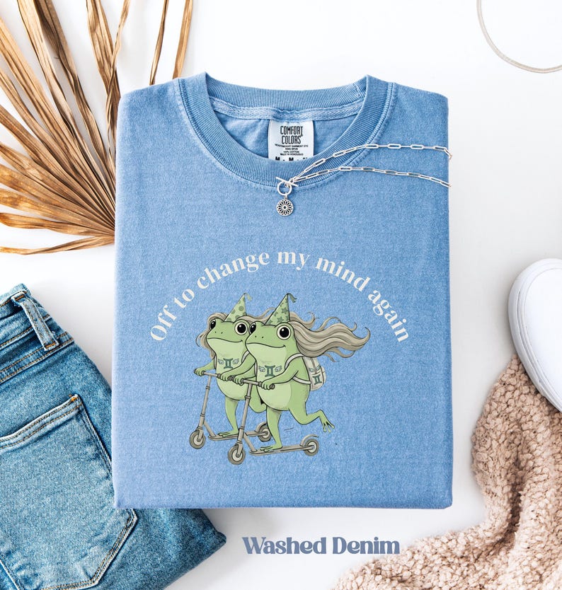 May include: Light blue t-shirt with the text "Off to change my mind again" and a graphic of two green frogs on scooters. The shirt is folded and has a Comfort Colors tag. The words "Washed Denim" are printed at the bottom.