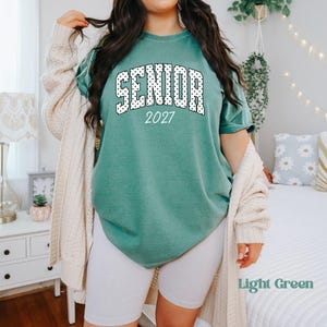 Peut inclure: Un t-shirt vert clair avec le mot "SENIOR" en lettres &agrave; pois blancs et l'ann&eacute;e "2027". Le t-shirt est associ&eacute; &agrave; un short blanc et un cardigan cr&egrave;me. Les mots "Light Green" sont dans le coin inf&eacute;rieur droit.