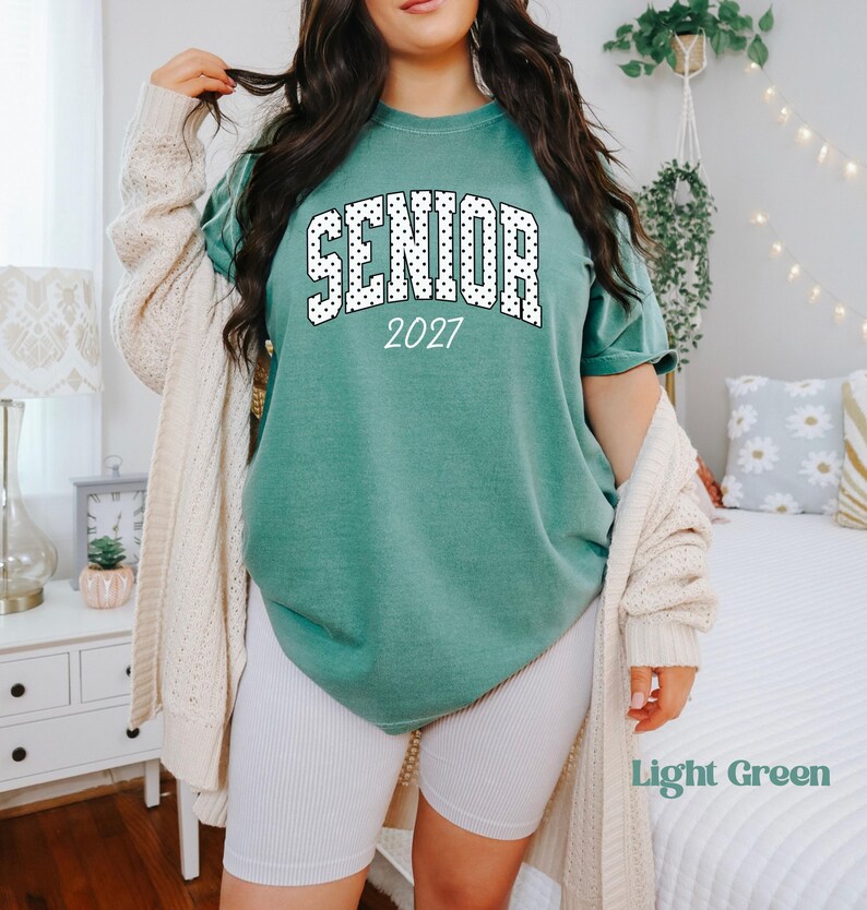 Peut inclure: Un t-shirt vert clair avec le mot "SENIOR" en lettres &agrave; pois blancs et l'ann&eacute;e "2027". Le t-shirt est associ&eacute; &agrave; un short blanc et un cardigan cr&egrave;me. Les mots "Light Green" sont dans le coin inf&eacute;rieur droit.