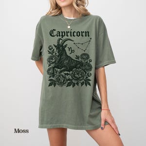 May include: Moss green t-shirt featuring a black Capricorn graphic. The design includes the word "Capricorn", a goat illustration, constellation, and floral elements. The shirt has short sleeves and a relaxed fit.