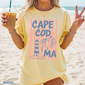Puede incluir: Una camiseta amarilla clara con las palabras "CAPE COD, MA" en azul, junto con rayas verticales rosas e ilustraciones de un faro, un lazo y una ostra. La palabra "Butter" está impresa en la esquina inferior izquierda.