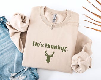 May include: A sand-colored sweatshirt with the text "He's Hunting." in green, along with a deer silhouette. The sweatshirt is folded, with the sleeves and a silver necklace visible. The word "Sand" is printed at the bottom.