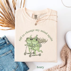 May include: Ivory-colored t-shirt with the text "Off to change my mind again." The shirt features a whimsical illustration of two green frogs riding scooters. A gold necklace and a pair of blue jeans are also visible.