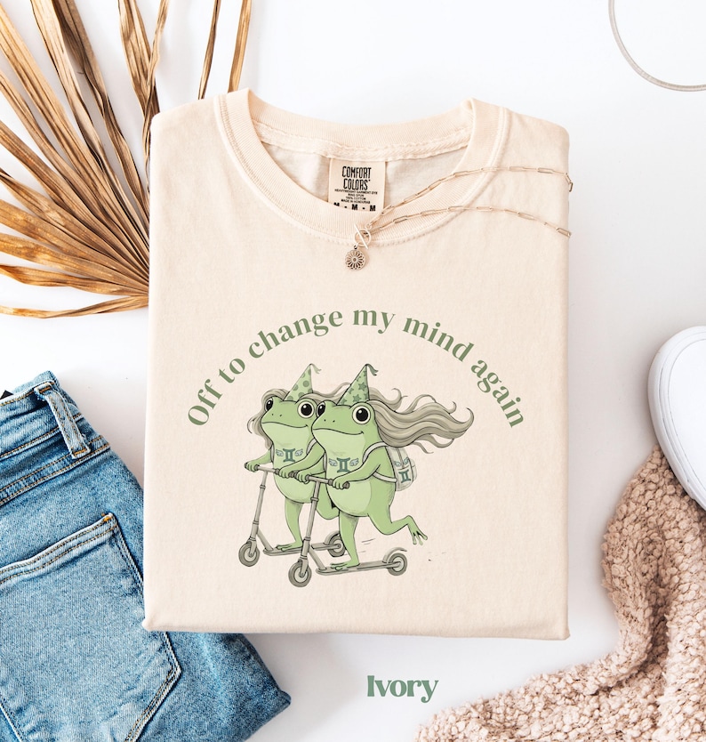 May include: Ivory-colored t-shirt with the text "Off to change my mind again." The shirt features a whimsical illustration of two green frogs riding scooters. A gold necklace and a pair of blue jeans are also visible.