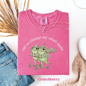 May include: A pink Comfort Colors t-shirt with the text "Off to change my mind again" encircling a graphic of two green frogs on scooters. The shirt also features the word "Crunchberry" and a silver necklace.