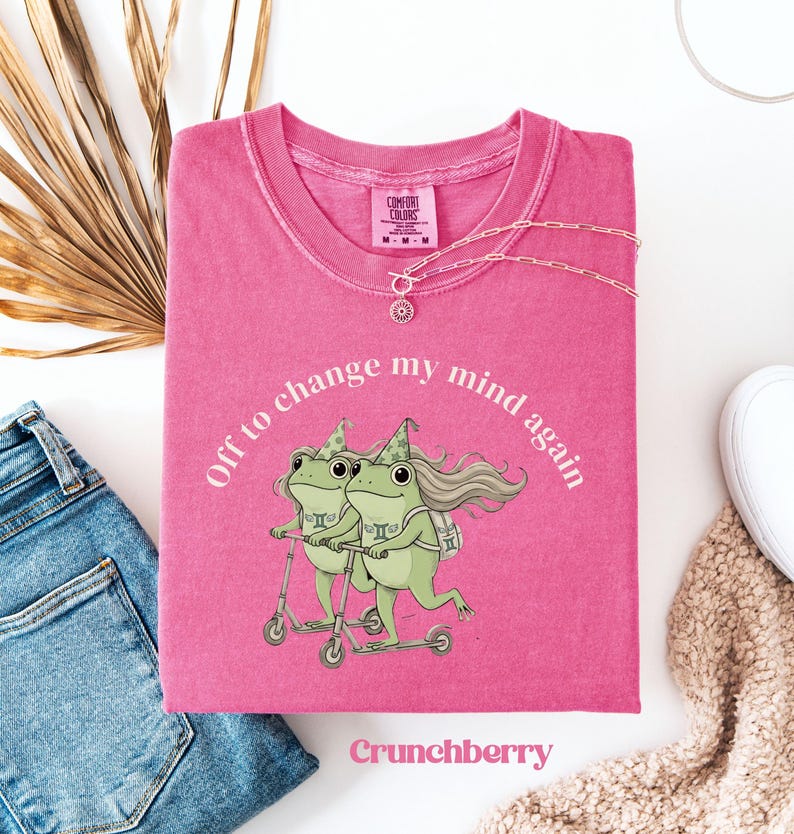 May include: A pink Comfort Colors t-shirt with the text "Off to change my mind again" encircling a graphic of two green frogs on scooters. The shirt also features the word "Crunchberry" and a silver necklace.
