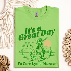 Puede incluir: Camiseta verde con un gráfico de una mujer montando un caballo y un perro corriendo en un paisaje desértico. El texto "It's a Great Day To Cure Lyme Disease" está impreso en la camiseta.
