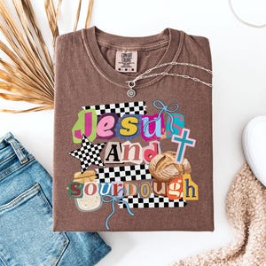 May include: Brown t-shirt with the words "Jesus And Sourdough" in a colourful, collage-style design. The shirt features a star, cross, and bread graphic. A silver necklace with a pendant is draped on the shirt, next to a pair of blue jeans.