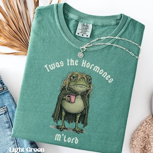 May include: A light green t-shirt featuring a graphic of a frog with long hair wearing a dark cape and holding a pink mug. The text above the frog reads Twas the Hormones and the text below reads M'lord. A silver chain necklace rests on the collar.
