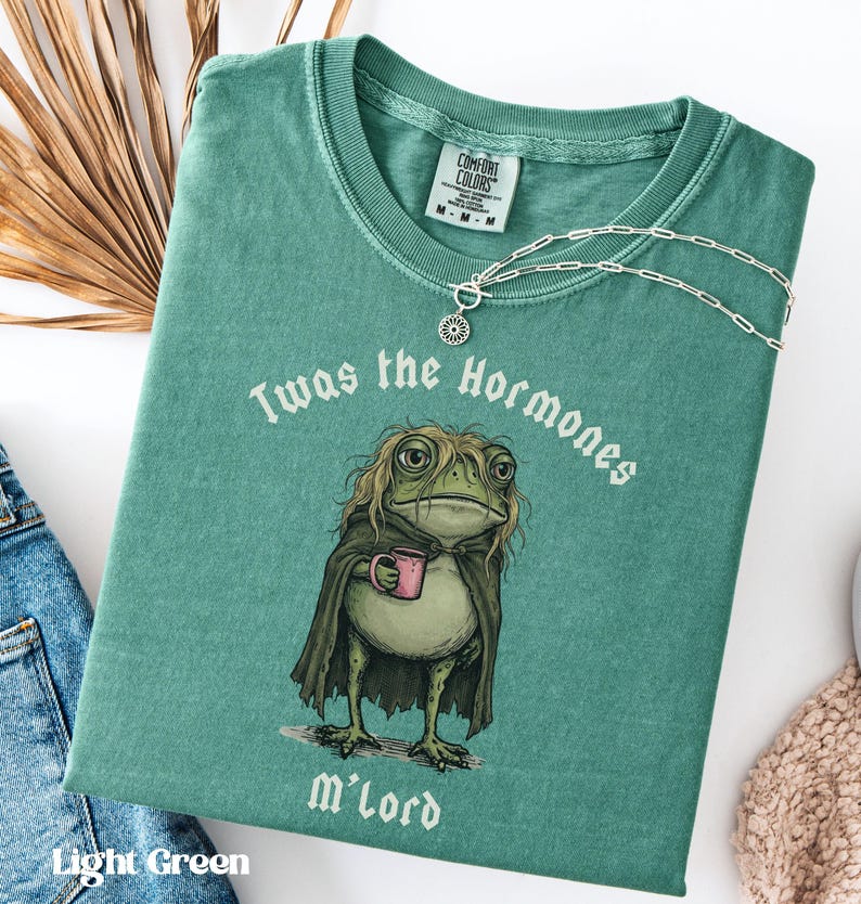 May include: A light green t-shirt featuring a graphic of a frog with long hair wearing a dark cape and holding a pink mug. The text above the frog reads Twas the Hormones and the text below reads M'lord. A silver chain necklace rests on the collar.