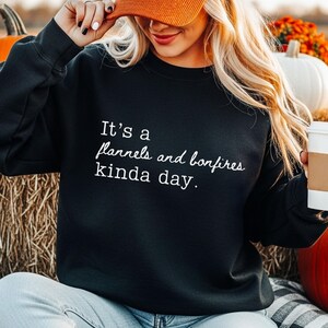 Puede incluir: Sudadera negra con el texto "It's a flannels and bonfires kinda day." La modelo lleva una gorra de pana naranja que dice "Pumpkin Spice". La sudadera se combina con vaqueros azules. La palabra "Black" est&aacute; en la parte inferior de la imagen.