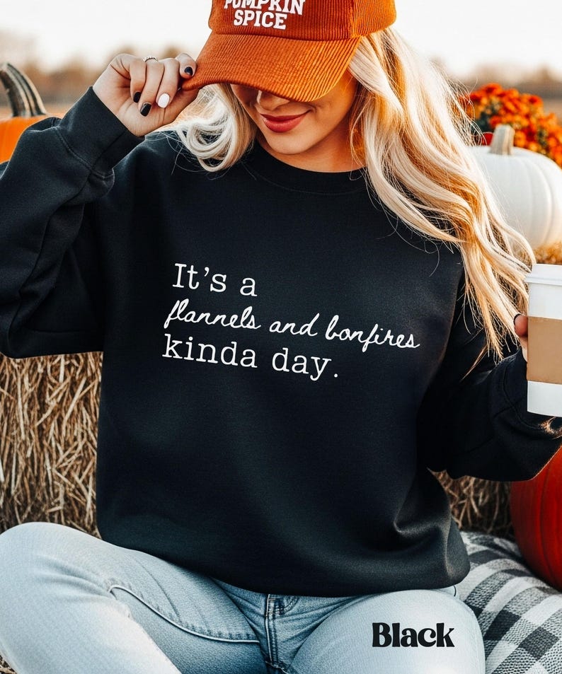 Puede incluir: Sudadera negra con el texto "It's a flannels and bonfires kinda day." La modelo lleva una gorra de pana naranja que dice "Pumpkin Spice". La sudadera se combina con vaqueros azules. La palabra "Black" est&aacute; en la parte inferior de la imagen.
