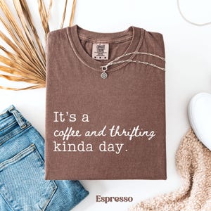 May include: Espresso-colored t-shirt with the text "It's a coffee and thrifting kinda day." A silver necklace with a pendant rests on the shirt. The shirt is folded, with a pair of blue jeans and a white shoe visible.