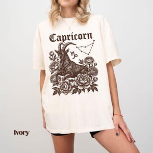 May include: Ivory-colored t-shirt featuring a Capricorn zodiac design. The graphic includes a detailed illustration of a goat, the word "Capricorn" in a decorative font, the Capricorn symbol, and a constellation outline, surrounded by floral elements.