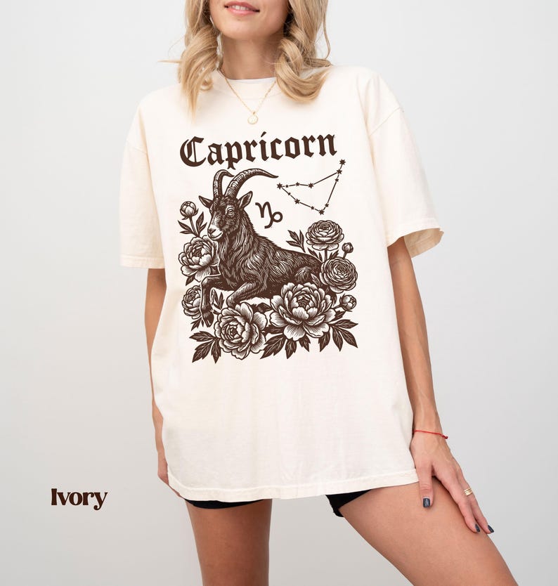May include: Ivory-colored t-shirt featuring a Capricorn zodiac design. The graphic includes a detailed illustration of a goat, the word "Capricorn" in a decorative font, the Capricorn symbol, and a constellation outline, surrounded by floral elements.