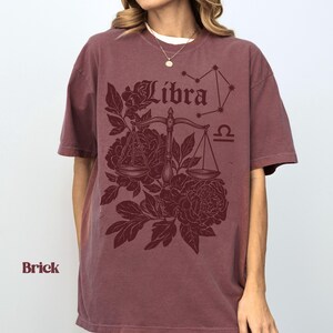 Op de afbeelding: Baksteenkleurig T-shirt met een Weegschaal-ontwerp. Het ontwerp bevat het woord "Libra", een weegschaal, bloemelementen en een sterrenbeeld. Het shirt heeft korte mouwen en een ronde hals.