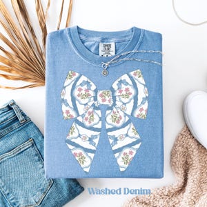 May include: A light blue Comfort Colors t-shirt with a large bow design. The bow features a white and blue pattern with pink floral accents. The words "Washed Denim" are printed at the bottom of the shirt. A silver necklace is draped across the shirt.