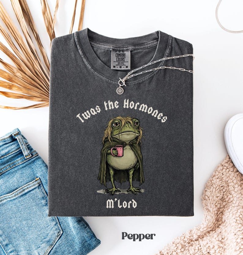 May include: A charcoal grey short-sleeve t-shirt featuring a graphic of a green frog with long hair wearing a cape and holding a pink mug. The text above the frog reads 'Twas the Hormones' and the text below the frog reads 'M'Lord' in white font.