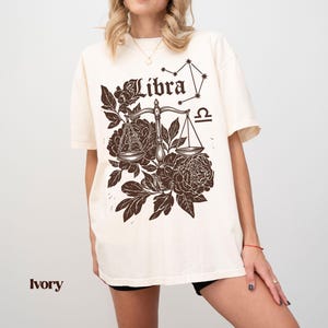 Op de afbeelding: Ivoor kleurig T-shirt met een Weegschaal sterrenbeeld ontwerp. Het ontwerp bevat een weegschaal, bloemelementen, het woord "Libra" en het Weegschaal symbool. Het shirt heeft korte mouwen en een relaxte pasvorm.