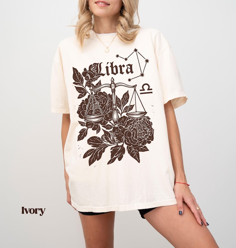 Op de afbeelding: Ivoor kleurig T-shirt met een Weegschaal sterrenbeeld ontwerp. Het ontwerp bevat een weegschaal, bloemelementen, het woord "Libra" en het Weegschaal symbool. Het shirt heeft korte mouwen en een relaxte pasvorm.