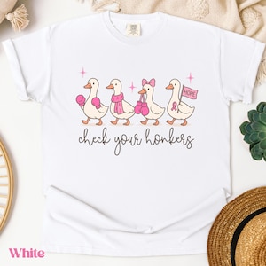 Pu&ograve; includere: T-shirt bianca con un'illustrazione di quattro anatre dei cartoni animati, ognuna adornata con accessori rosa come guantoni da boxe e sciarpe. Il testo "check your honkers" &egrave; scritto sotto, e una bandiera con la parola "HOPE" &egrave; inclusa.