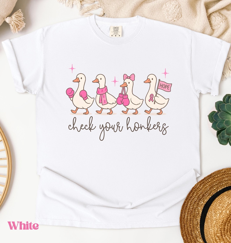 Pu&ograve; includere: T-shirt bianca con un'illustrazione di quattro anatre dei cartoni animati, ognuna adornata con accessori rosa come guantoni da boxe e sciarpe. Il testo "check your honkers" &egrave; scritto sotto, e una bandiera con la parola "HOPE" &egrave; inclusa.