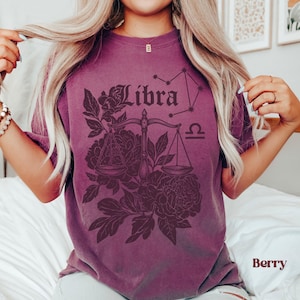 Op de afbeelding: Bessenkleurig T-shirt met een Weegschaal sterrenbeeld ontwerp. Het ontwerp bevat het woord "Libra", een weegschaal, een sterrenbeeld en bloemmotieven. Het shirt is effen van kleur met het woord "Berry" rechtsonder gedrukt.