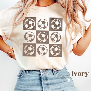 May include: Ivory t-shirt with a brown and white soccer ball pattern. The shirt has a crew neck and short sleeves.