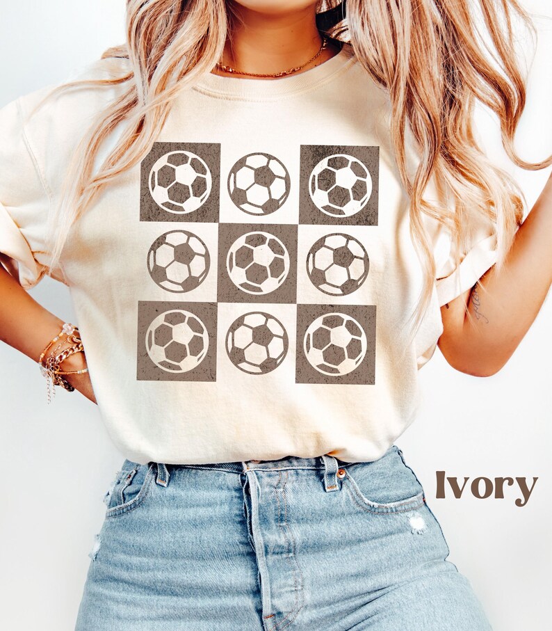 May include: Ivory t-shirt with a brown and white soccer ball pattern. The shirt has a crew neck and short sleeves.