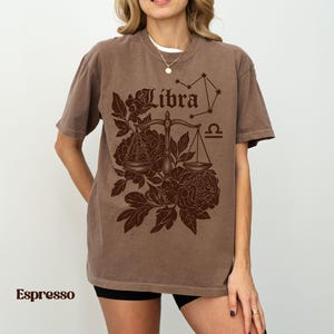 Op de afbeelding: Espresso-kleurig T-shirt met een Weegschaal sterrenbeeld ontwerp. Het ontwerp bevat het woord "Libra", een weegschaal, bloemelementen en een sterrenbeeldgrafiek. Het shirt heeft korte mouwen en een relaxte pasvorm.