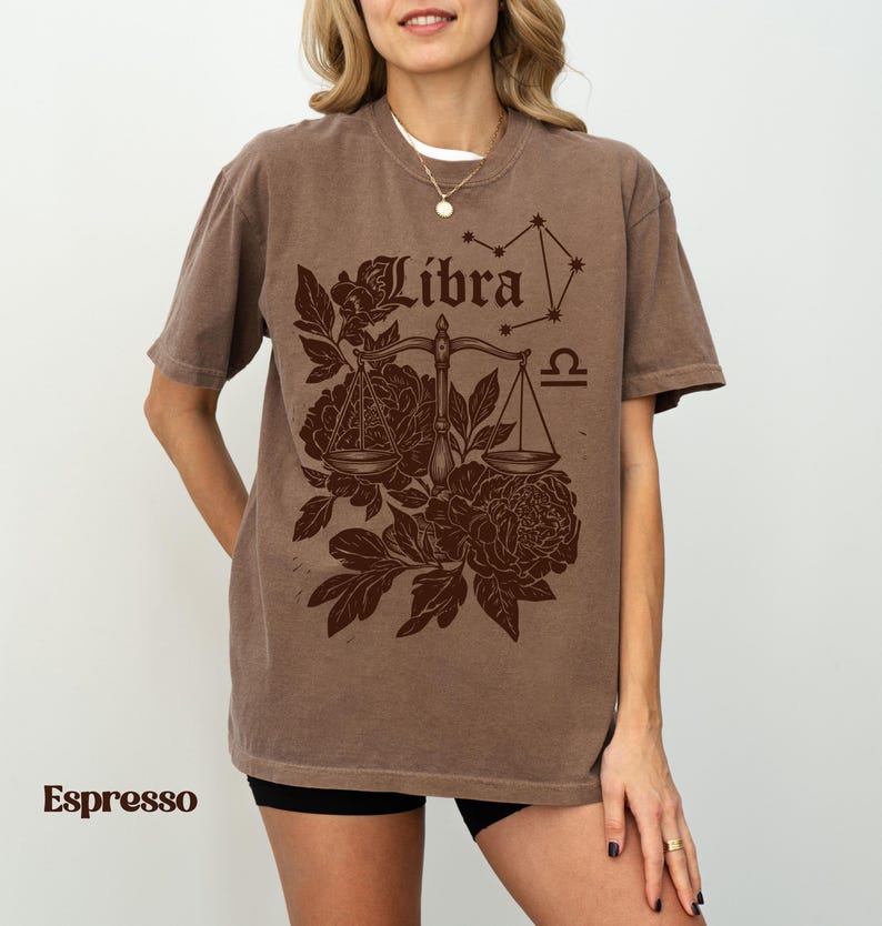 Op de afbeelding: Espresso-kleurig T-shirt met een Weegschaal sterrenbeeld ontwerp. Het ontwerp bevat het woord "Libra", een weegschaal, bloemelementen en een sterrenbeeldgrafiek. Het shirt heeft korte mouwen en een relaxte pasvorm.