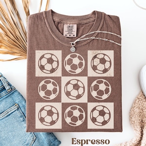 May include: Espresso brown t-shirt with a vintage-style design of nine soccer balls in a grid pattern. The shirt has a Comfort Colors tag.