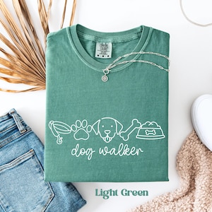 Könnte beinhalten: Hellgrünes T-Shirt mit weißer Grafik von Utensilien für Hundeauslauf und dem Text "dog walker". Eine silberne Halskette mit Anhänger liegt auf dem Shirt. Das Shirt liegt gefaltet neben einer blauen Jeans und einem weißen Schuh.