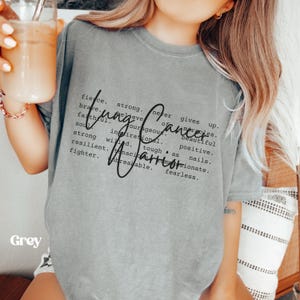 May include: Gray t-shirt with the text "Lung Cancer Warrior" defined as: fierce, strong, never gives up, believe in miracles, brave, courageous, beautiful, faithful, inspirational, positive, strong willed, tough as nails, resilient, tenacious, passionate, fighter, unbreakable, fearless.