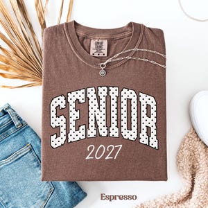 Peut inclure: T-shirt couleur expresso avec le mot "SENIOR" en blanc, lettres pointill&eacute;es et l'ann&eacute;e "2027". Le t-shirt est pr&eacute;sent&eacute; avec un jean bleu, un collier argent&eacute; et une chaussure blanche. Le t-shirt est de Comfort Colors.