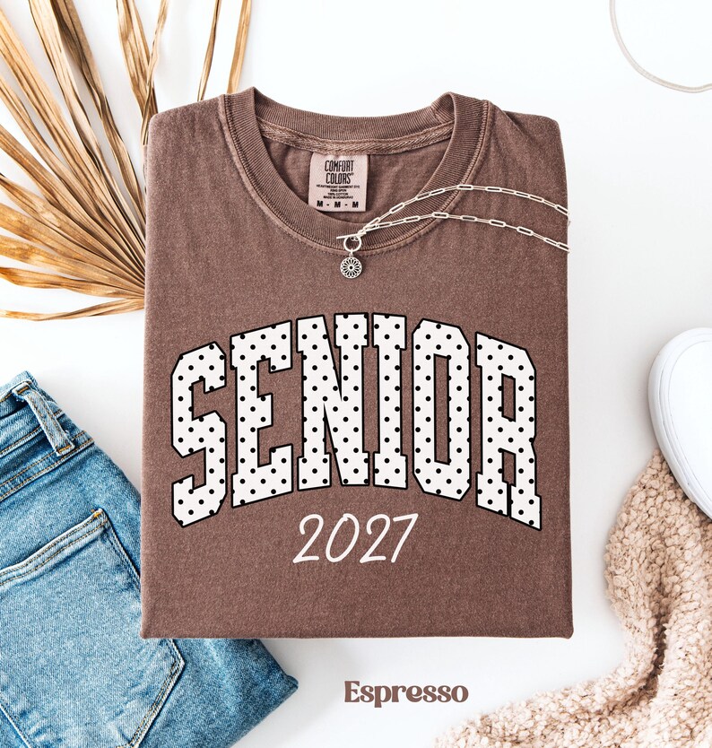 Peut inclure: T-shirt couleur expresso avec le mot "SENIOR" en blanc, lettres pointill&eacute;es et l'ann&eacute;e "2027". Le t-shirt est pr&eacute;sent&eacute; avec un jean bleu, un collier argent&eacute; et une chaussure blanche. Le t-shirt est de Comfort Colors.