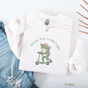 Può includere: Felpa bianca con grafica di una rana verde con cappello su uno scooter. Il testo "On my way to the Cape" è stampato sopra la rana. La parola "White" è stampata in basso a destra.