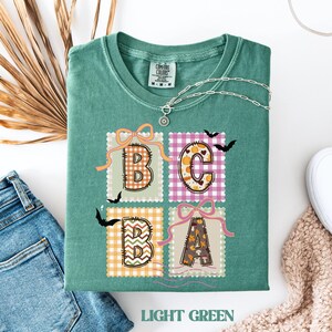 Puede incluir: Camiseta verde claro con el texto "BCBA" en un diseño cuadrado. Cada letra tiene un patrón diferente, con murciélagos y lazos. La camiseta está doblada con un collar plateado y unos vaqueros.
