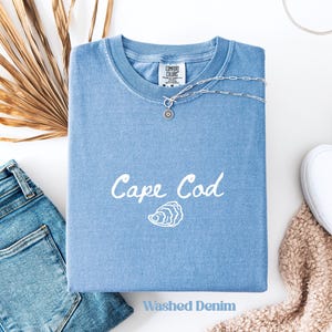 Puede incluir: Una camiseta azul claro "Washed Denim" con la inscripción "Cape Cod" y un gráfico de ostra. Un collar plateado con un colgante descansa sobre la camisa. La camiseta está doblada con unos vaqueros azules y un zapato blanco cerca.